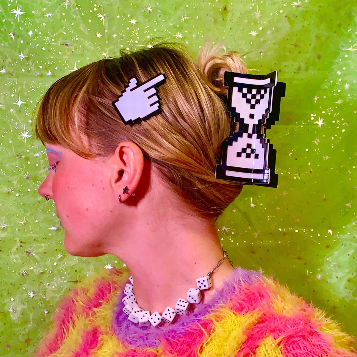 A side-profile portrait of a woman with blonde hair in a bun, wearing two pixel-art hair clips: a pointing hand clip and an hourglass clip. 