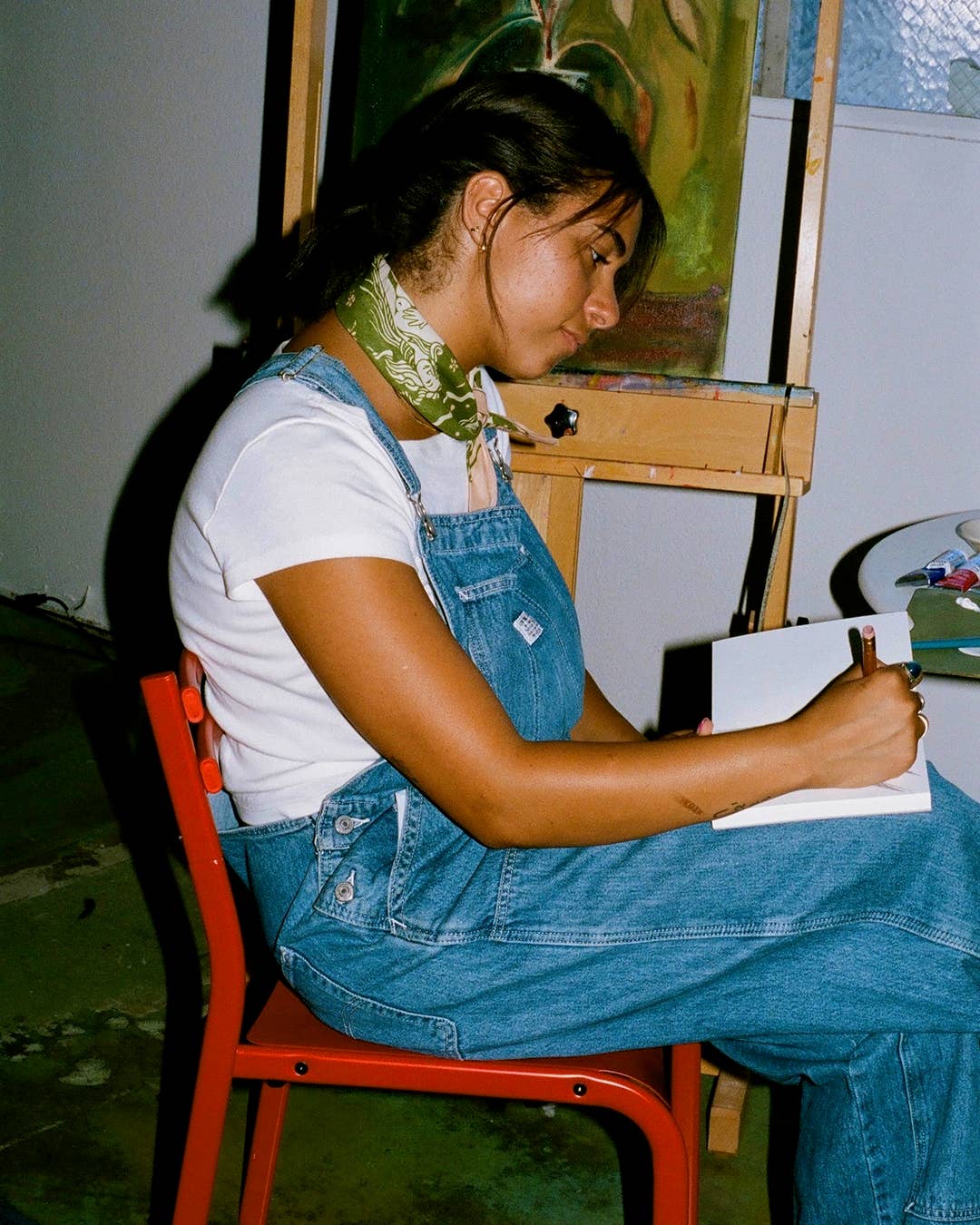 Person wearing green bandana as necklace sitting on a red chair drawing at an easel with a painting in the background.