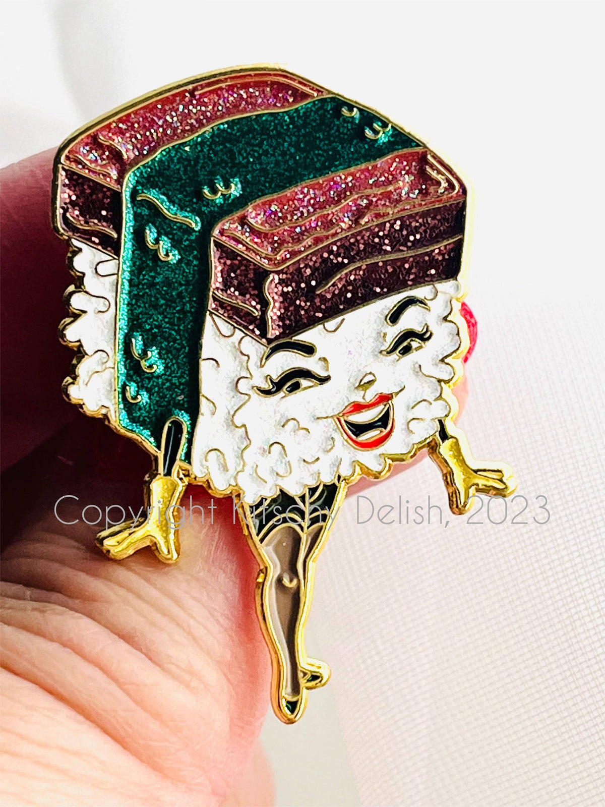 A close-up of a hand holding a colorful enamel pin shaped like a smiling sushi character with glittery green and red toppings, white rice, and gold outlines, against a light background.