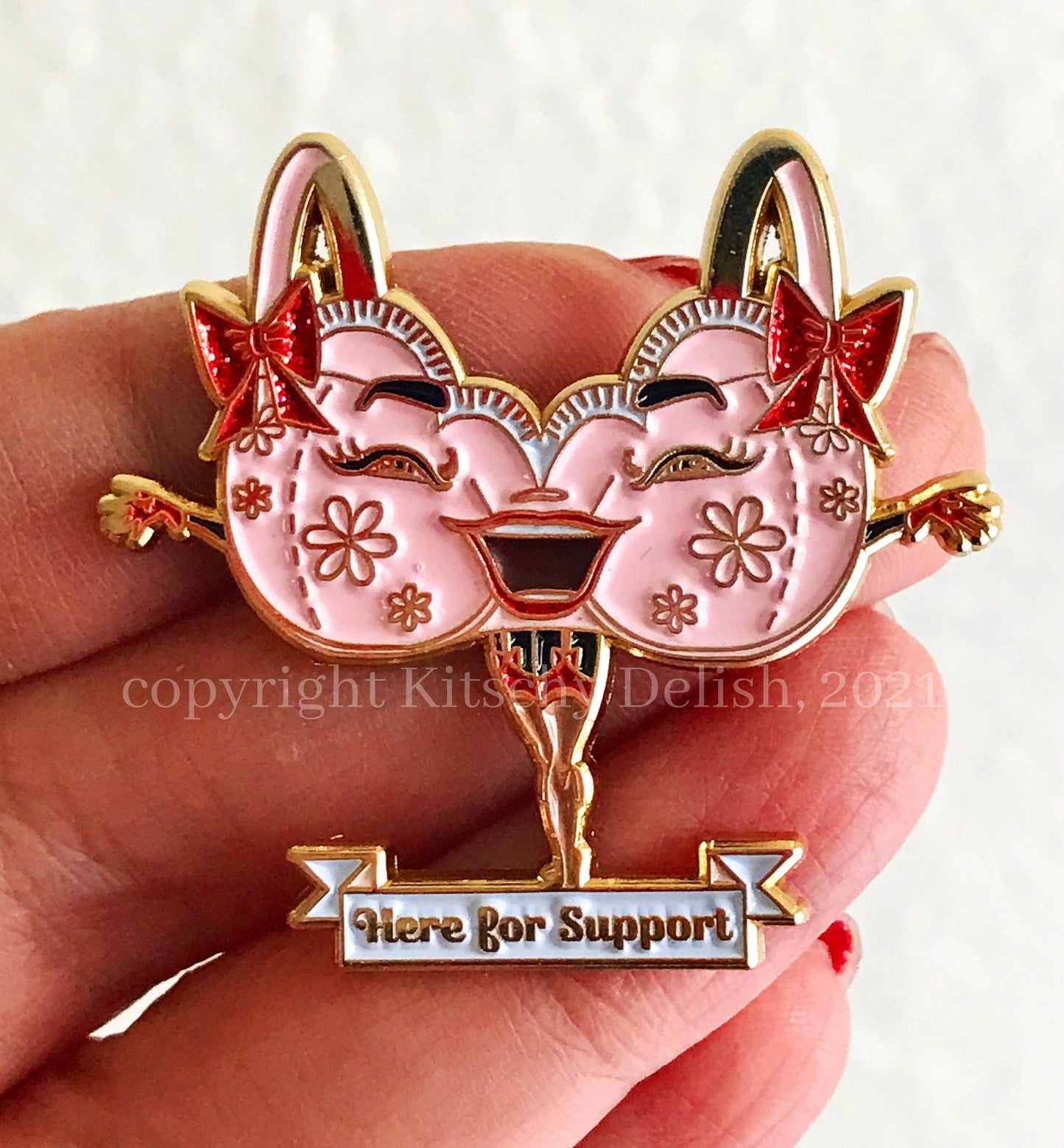 A close-up of a hand holding a decorative enamel pin shaped like two smiling pink faces with gold outlines, red bows, floral details, and a banner reading 'Here for Support' at the bottom.