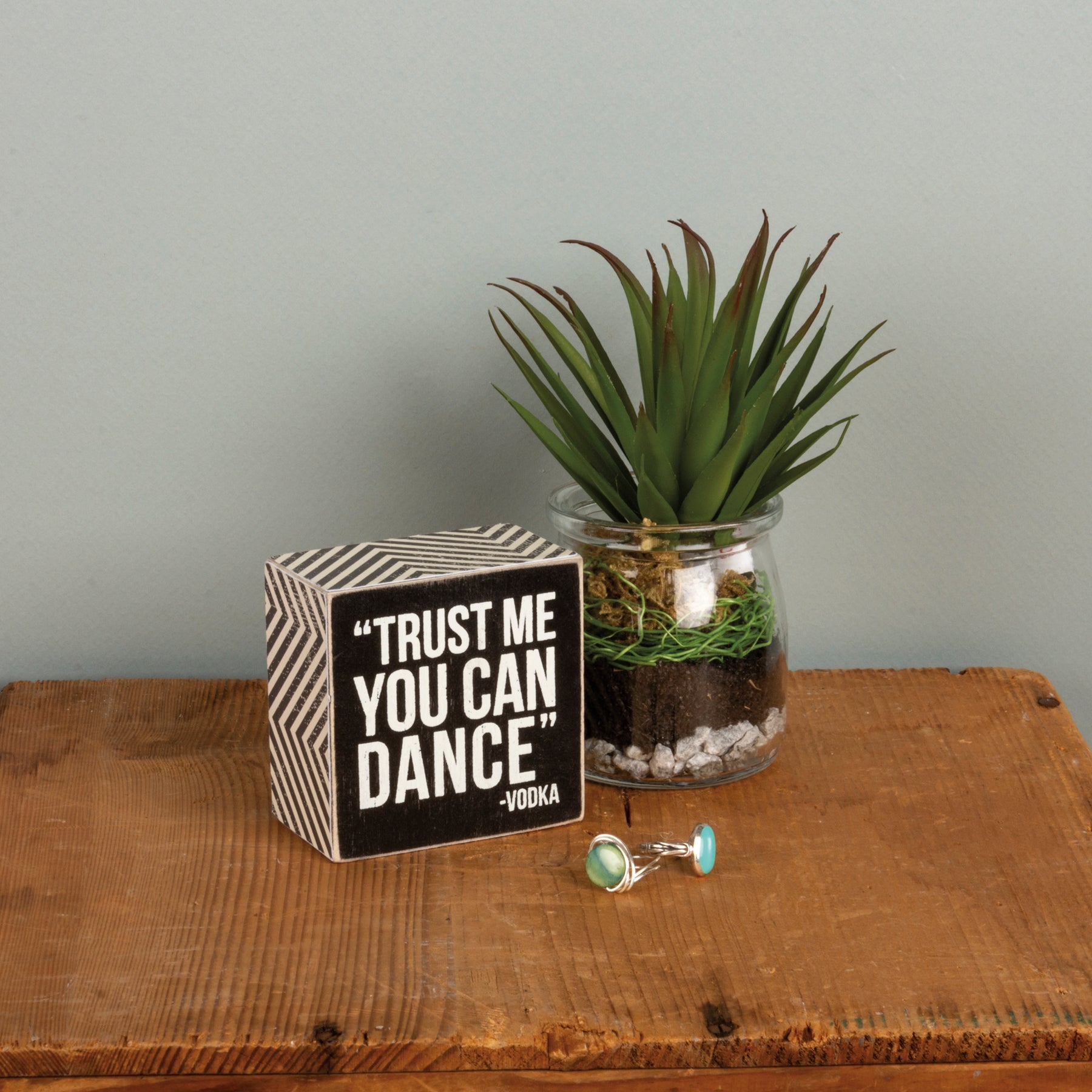 A classic black and white wooden box sign lending a distressed "'Trust Me You Can Dance' - Vodka" sentiment with zigzag patterned trim styled on wooden table.